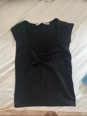 Svart topp med rosett från 157 - Säljer en svart topp från 157 med liten rosett och rynkdetalj framtill. Toppen har djup v-ringning och spetskant längs urringningen. Perfekt att styla med jeans eller kjol för en trendig look.