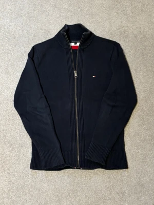 Full zip Tommy Hilfiger - Snygg marinblå ziptröja från Tommy Hilfiger med hög krage och diskret logga på bröstet. Tröjan är ribbstickad med breda muddar vid ärmslut och nederkant. Perfekt för lager på lager och enkel att matcha med olika outfits.