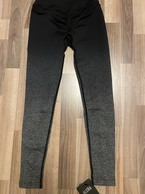 Svarta/grå ombre leggings från Alera - Leggings från Alera i snyggt svart till grått ombre-mönster. Hög midja och scrunch-detalj bak för extra shaping. Tillverkade i stretchigt material som sitter tajt och bekvämt. Perfekta för dig som vill ha både stil och komfort.