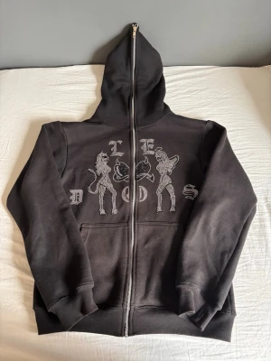 Svart Rhinestone hoodie  - Cool svart hoodie från DOS med hel dragkedja och huva. På framsidan finns ett unikt motiv med två figurer och text i glittrande stenar. Hoodien har ribbade muddar och fickor framtill. Perfekt för dig som gillar streetwear och vill sticka ut.