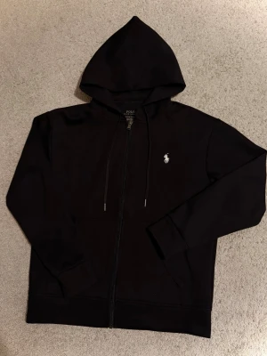 Svart hoodie från Polo Ralph Lauren - Klassisk svart hoodie med dragkedja från Polo Ralph Lauren. Har huva med dragsko, två fickor framtill och den ikoniska vita logon broderad på bröstet. Perfekt för en chill och stilren look.