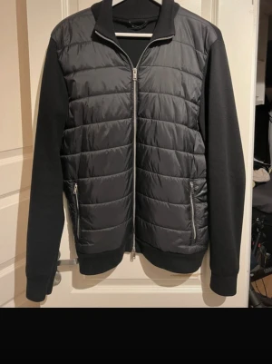 Svart quiltad cardiganjacka - Snygg svart cardiganjacka med quiltad framsida och mjuka ärmar. Jackan har dragkedja framtill, två fickor med dragkedja och ribbade muddar. Perfekt för lager-på-lager och en clean look.