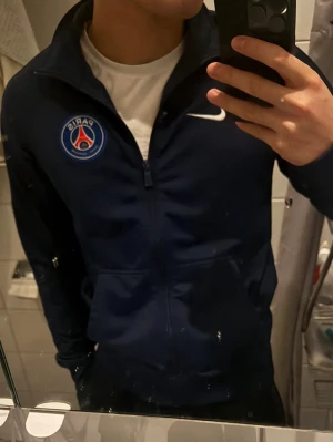 Mörkblå PSG zip-tröja från Nike - Snygg mörkblå zip-tröja från Nike med Paris Saint-Germain-logga på bröstet och vit Nike swoosh. Tröjan har hög krage och dragkedja framtill, perfekt för dig som gillar sportig stil och vill representera PSG.