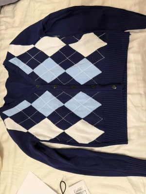 Mörkblå argyle kofta med knappar - Snygg mörkblå kofta med klassiskt argylemönster i ljusblått och vitt. Koftan har v-ringning, långa ärmar och svarta knappar framtill. Ribbstickade muddar vid ärmslut och nederkant. Perfekt för en preppy look.