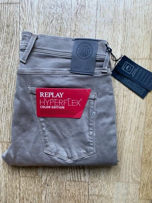 Beige Replay Hyperflex jeans - Säljer ett par snygga beige Replay Hyperflex jeans i stretchigt material. Modellen har klassisk femficksdesign, normal midja och smal passform. Perfekta för dig som vill ha både stil och komfort. Märkta med Replay-logga och har coola detaljer.