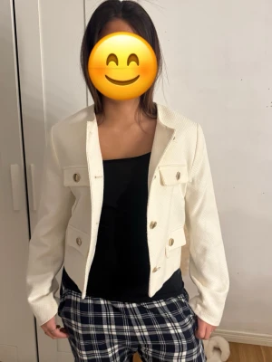 Vit cardiganjacka med guldknappar - Stilren vit cardiganjacka med strukturerat tyg och dekorativa guldknappar framtill. Jackan har två bröstfickor och två fickor vid midjan, samt en rak, kortare passform. Perfekt att slänga över en topp för en chic look.