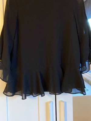 Svart volangblus  - Snygg svart blus med volangkant nedtill och vid passform. Blusen har rund halsringning och långa, lätt transparenta ärmar. Perfekt för dig som gillar en luftig och trendig stil.