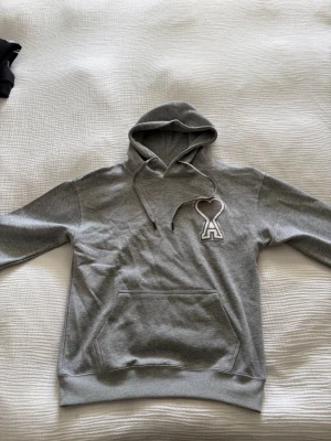 Ami Paris hoodie - Snygg grå hoodie från AMI Paris med stor broderad hjärtlogga på bröstet. Klassisk känguruficka framtill, ribbade muddar och dragsko i huvan. Tillverkad i mjuk bomullsblandning som känns skön mot huden. Perfekt för en avslappnad och trendig look.