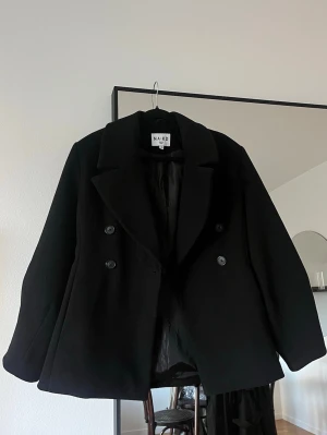 Svart kort kappa från NA-KD - Stilren svart kappa från NA-KD med dubbelknäppning och bred krage. Sitter snyggt oversized på mig som är en xs och 164 lång. 🖤 mycket bra skick!