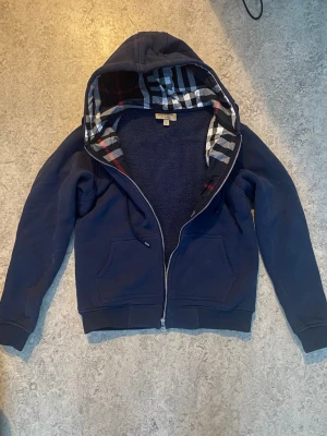Mörkblå hoodie från Burberry - Snygg mörkblå hoodie från Burberry med dragkedja och klassiskt rutigt foder i huvan. Hoodien har två fickor framtill och ribbade muddar vid ärmar och nederkant. Perfekt för en chill och stilren look.