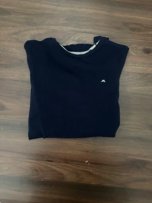 Mörkblå stickad tröja J.Lindeberg - Mörkblå stickad tröja från J.Lindeberg med rund halsringning och diskret logga på bröstet. Tröjan har långa ärmar och ribbade muddar vid ärmslut och nederkant. Perfekt för lager på lager och stilren look storlek m men passar s.