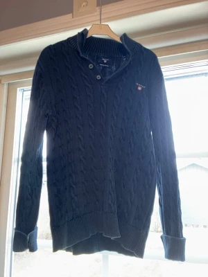 Mörkblå kabelstickad tröja Gant - Mörkblå kabelstickad tröja från Gant med tre knappar vid kragen och liten broderad logga på bröstet. Tröjan har lång ärm och en klassisk krage. Perfekt för en stilren och avslappnad look. Det är bara att höra av sig vid frågor/funderingar! Nypris runt 1500!