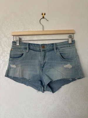Ljusblå jeansshorts  - Säljer ett par ljusblå jeansshorts med rå kant och slitna detaljer framtill. Klassisk femficksmodell med knapp och dragkedja. Perfekta till sommaren och har en avslappnad passform. Är lågmidjan och är i storlek xs
