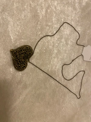 Halsband med antikt hjärthänge - Snyggt halsband med ett stort hjärthänge i antikguld. Hänget har ett detaljerat, genombrutet mönster som ger en vintagekänsla. Kedjan är tunn och matchar hängets färg. Perfekt accessoar för dig som gillar unika smycken med romantisk touch. Copper färg 
