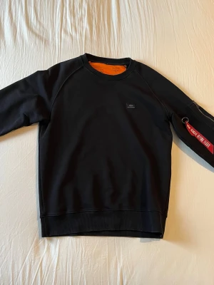 Svart sweatshirt från Alpha Industries - Cool svart sweatshirt från Alpha Industries i mycket bra skick. Unik detalj med dragkedjeficka på ärmen och röd 'Remove Before Flight'-tag. Nypris: 1499kr, Mitt pris: 699kr / Skriv vid frågor 🤝