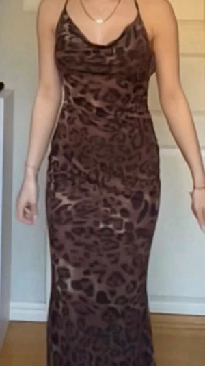 Brun leopardmönstrad långklänning - Snygg långklänning med leopardmönster i bruna toner. Klänningen har tunna axelband som korsas i ryggen och en smickrande, figurnära passform. Materialet är stretchigt och lätt, perfekt för dig som vill sticka ut. (Ursäkta bildkvaliteten, screenshots från en video)