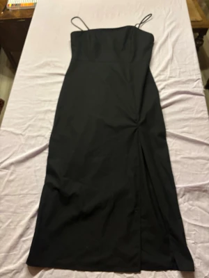 Svart långklänning med tunna axelband och slits  - Stilren svart långklänning med tunna axelband slits och figurnära passform. Klänningen har en enkel, elegant design utan synliga detaljer eller mönster, vilket gör den lätt att styla. Perfekt för dig som gillar minimalistisk och klassisk stil.