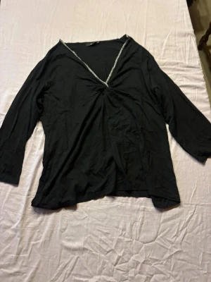 Svart v-ringad topp med lång ärm - Svart topp med v-ringning och dekorativ kant i halsen. Toppen har långa ärmar och en lätt rynkning framtill vid ringningen. Passar perfekt till jeans eller kjol för en enkel men snygg look.