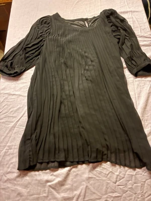 Svart plisserad klänning med puffärm - Snygg svart klänning med plisserat tyg och puffiga trekvartsärmar. Klänningen har rund halsringning och en liten öppning bak i nacken. Tillverkad i ett lätt, flowy material som ger en elegant look.