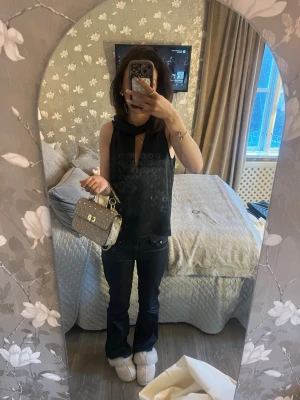 Svart ärmlös topp med scarfdetalj - Snygg svart ärmlös topp med scarfdetalj runt halsen och en liten v-ringning framtill. Toppen har en elegant och stilren look, perfekt för att lyfta vilken outfit som helst. Materialet ser ut att vara mjukt och följsamt, troligen polyester.