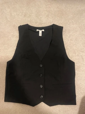 Svart kostymväst från H&M - Stilren svart kostymväst från H&M med fyra knappar framtill och två dekorativa fickor. Västen har en klassisk V-ringning och är perfekt att matcha med kostymbyxor för en dressad look. Materialet är slätt och ger en snygg siluett.