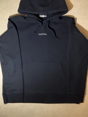Svart hoodie från Valentino - Snygg svart hoodie från Valentino med  logga på bröstet. Klassisk känguruficka framtill, justerbar huva med dragsko och ribbade muddar. Tillverkad i mjukt material som känns skönt mot huden. Perfekt för en stilren och avslappnad look.