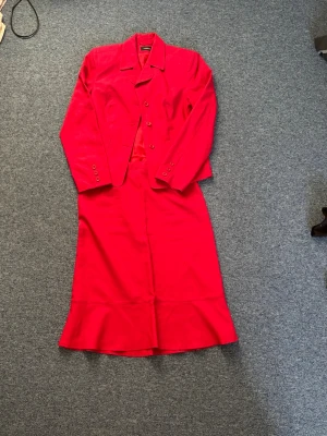 Röd kavaj och kjol från Lindex - Stilren röd kavaj och kjol från Lindex med klassisk krage och tre knappar framtill. Kavajen har figurnära passform, långa ärmar och dekorativa knappar vid ärmsluten. Den tillhörande är i samma material och färg med dragkedja. Settet har en snygg färg som ger en professionell och modern look. Oanvänd utan defekter, fläckarna är endast damm då den inte kommit till användning. 