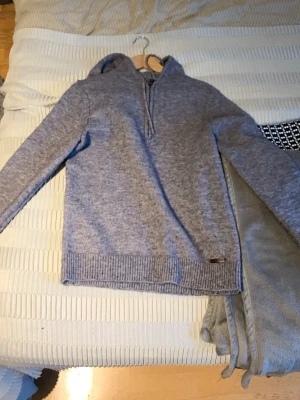 Grå stickad hoodie från Pier One - Mysig grå stickad hoodie från Pier One med ribbade muddar och huva. Perfekt för kyliga dagar och enkel att matcha med jeans eller mjukisbyxor. Klassisk modell med lång ärm och diskret logga vid nederkanten.