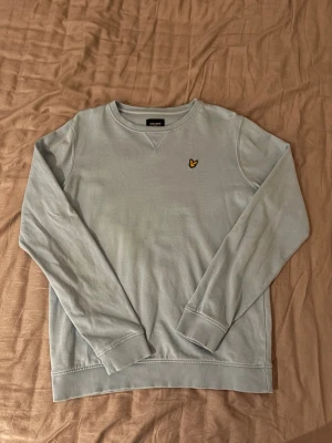Ljusblå Lyle & Scott tröja - Snygg ljusblå sweatshirt från Lyle & Scott med klassisk rund halsringning och ribbade muddar. Tröjan har det ikoniska gula örnlogot på bröstet och är tillverkad i mjukt bomullsmaterial. Perfekt för en avslappnad och stilren look.