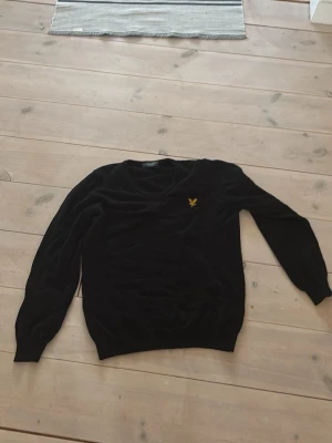Svart stickad tröja från Lyle & Scott - Snygg svart stickad tröja från Lyle & Scott med klassisk gul broderad örn på bröstet. Tröjan har V-ringning och långa ärmar. . Materialet är mjukt och följsamt, vilket gör den riktigt skön att bära.