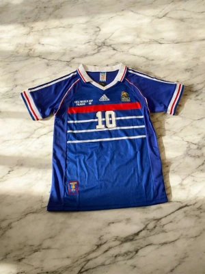 Blå Frankrike fotbollströja Adidas - Säljer en blå fotbollströja från Adidas med Frankrikes landslagslogga och nummer 10, inspirerad av Zidane från VM 1998. Tröjan har vita och röda detaljer, korta ärmar och klassisk v-ringad krage. Perfekt för fotbollsfans!
