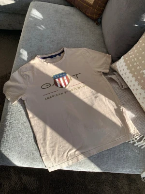 Beige GANT t-shirt med tryck - Snygg beige t-shirt från GANT med stort tryck framtill som visar ett sköldmärke med rödvita ränder och blått fält samt texten 'GANT American Sportswear'. Klassisk rund hals och korta ärmar. Perfekt till jeans eller shorts. Storlek 158/164, 13-14 år.
