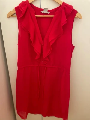 Röd ärmlös blus med volang - Säljer en röd ärmlös blus med volangdetaljer. Blusen har knappar framtill och ett tunt, lätt material som ger ett luftigt intryck. Perfekt för dig som gillar volanger och fina sommarkläder 💝