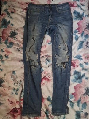 Blå slitna skinny jeans med hål - Säljer ett par blå skinny jeans med stora slitningar och hål på båda benen från Hollister. Jeansen har klassisk femficksdesign och normal midja. Perfekta för en avslappnad och trendig streetstil. Materialet är denim i bomull.