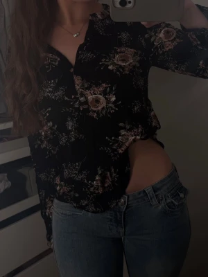 Svart blommig blus med knappar - Snygg svart blus med romantiskt blommönster i rosa och beige toner. Blusen har knappar framtill och långa ärmar. Perfekt för dig som vill ha en feminin och trendig look. Materialet ser ut att vara lätt och skönt, troligen viskos eller polyester.