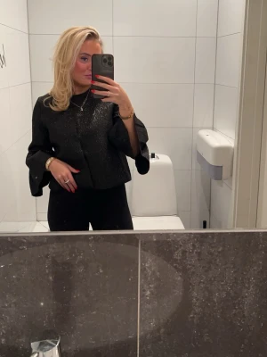 Svart glittrig topp med vida ärmar - Snygg svart topp med glittrigt tyg och vida trekvartsärmar. Toppen har en rak passform och är perfekt för dig som vill sticka ut lite extra. Matcha med svarta byxor för en clean look.