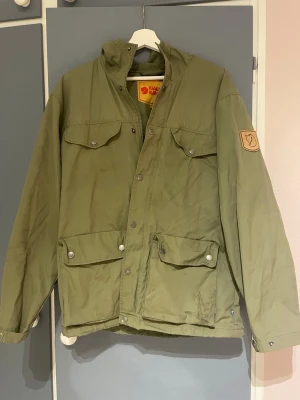 Grön vindjacka från Fjällräven - Stilren grön vindjacka från Fjällräven med fyra framfickor, tryckknappar och huva. Jackan har klassisk patch med logga på ärmen och är tillverkad i slitstarkt material som passar perfekt för utomhusaktiviteter. Enkel design med hög krage och justerbara detaljer.Ärm: 61cm Armhåla och nedåt: 41cm Krage och nedåt: 62cm Full längd från ryggen: 71cm