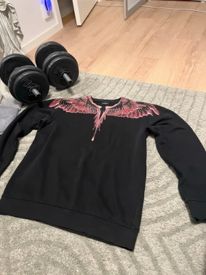 Svart sweatshirt med vingtryck - Säljer en svart sweatshirt från Marcelo Burlon med ikoniskt vingmönster i rött och rosa över axlar och bröst. Tröjan har rund halsringning och långa ärmar. Perfekt för dig som vill sticka ut med en unik design.