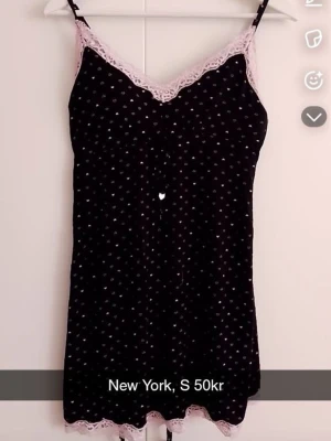 Svart nattlinne med spetsdetaljer - Sött svart nattlinne från New York med tunna axelband och små vita prickar. Dekorerat med ljusrosa spets längs urringning och nederkant för en romantisk touch. Perfekt för mysiga kvällar hemma.