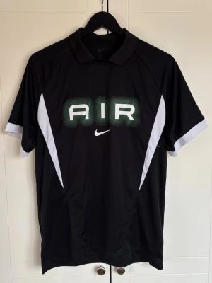 Svart Nike AIR t-shirt med tryck - Snygg svart t-shirt från Nike med stort AIR-tryck i grönt och vitt framtill samt klassisk Nike-logga. T-shirten har vita detaljer på sidorna och är kortärmad med krage. Perfekt för dig som gillar sportig och stilren design.