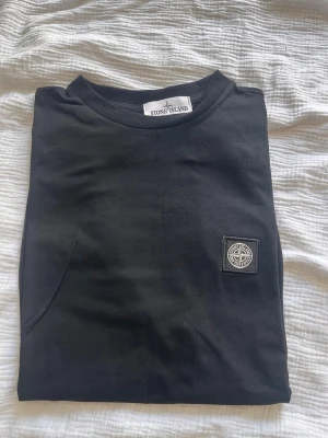 Svart Stone Island t-shirt - Svart t-shirt från Stone Island med klassisk rund hals och korta ärmar. T-shirten har en stilren design med Stone Island-logga på bröstet. Tillverkad i mjuk bomull som känns skön mot huden. Perfekt för en clean och trendig look.