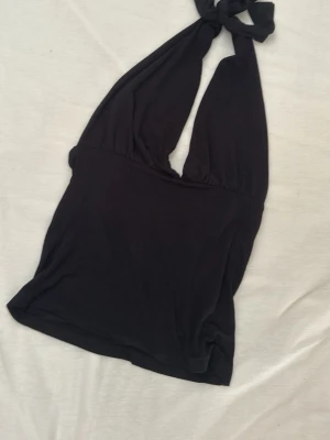 Svart halterneck topp med knyt - Snygg svart halterneck topp med knyt i nacken och djup v-ringning. Toppen är ärmlös och har en tight passform som framhäver figuren. Perfekt att styla med jeans eller kjol för en trendig look.