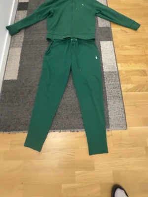 Grönt tracksuit från Polo Ralph Lauren - Mysigt grönt loungewear-set från Polo Ralph Lauren med dragkedja på tröjan, huva och fickor framtill. Byxorna har dragsko i midjan och smala ben. Liten vit logga broderad på både tröja och byxa. Perfekt för chill dagar hemma passar även S och xs