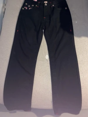Svarta True Religion jeans med röd detalj - Säljer ett par svarta jeans från True Religion med raka ben och röda kontrastsömmar. Jeansen har klassiska fickor fram och bak, samt de ikoniska True Religion-detaljerna på bakfickorna. Materialet är denim med stretch för extra komfort. Finns även påsen till och prislapp pris kan diskuteras.