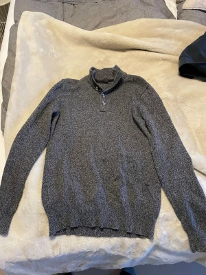 Grå stickad tröja  - Säljer en snygg gråmelerad stickad tröja. Tröjan har hög krage med knappslå och ribbade muddar vid ärmslut och nederkant. Perfekt för dig som gillar stilrena och klassiska plagg med en modern touch.