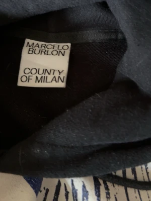 Svart hoodie Marcelo Burlon med vingar - Svart hoodie från Marcelo Burlon County of Milan med ikoniskt blått vingtryck över axlarna och ryggen. Hoodien har huva och ribbade muddar. Perfekt för dig som vill sticka ut med en streetwear-look.