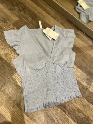 Ljusblå omlottopp med volangärm - Supersöt ljusblå omlottopp från H&M med volangärmar och smockad midja. Toppen har v-ringning och är tillverkad i ett lätt, skönt material som passar perfekt till jeans eller kjol. Enkel att styla för en trendig look.