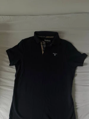 Svart pikétröja från Barbour - Klassisk svart pikétröja från Barbour med korta ärmar och krage. Tröjan har ett diskret rutigt tyg på insidan av knäppningen och Barbour-logga broderad på bröstet. Perfekt för en stilren och avslappnad look.