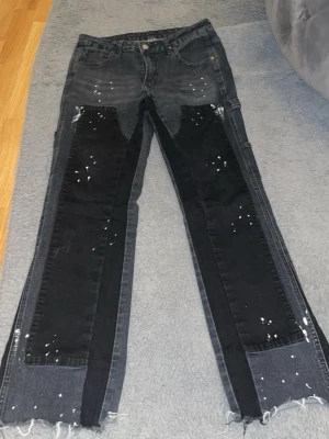 Svarta patchade jeans med råa kanter - Unika svarta jeans med kontrasterande svarta patchar och vita färgstänk. Raka ben med råa, ofållade kanter för en edgy look. Jeansen har fem fickor och cool streetstyle-vibe. Perfekta för dig som vill sticka ut med din outfit.