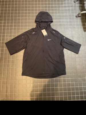 Svart Nike Miller - Svart vindjacka från Nike med huva och dragkedja framtill. Jackan har trekvartsärmar, reflekterande detaljer och en minimalistisk design. Perfekt för blåsiga dagar och sportiga outfits. Tillverkad i lätt och funktionellt material.